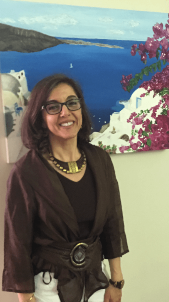 A woman with glasses and a brown blouse smiling in front of a painting of a seaside town