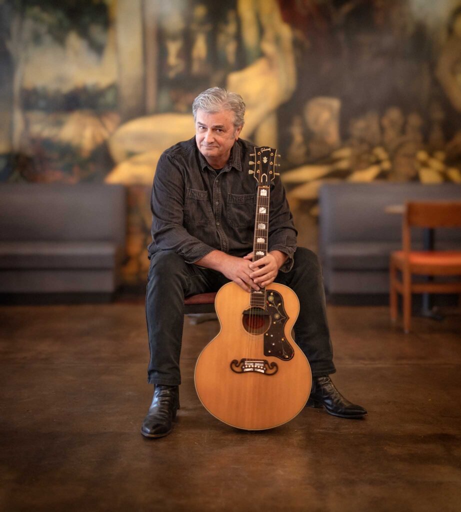 A man sits on a chair in a café, holding an acoustic guitar, with a blurred mural in the background.