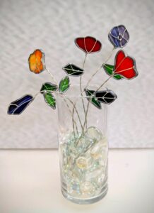 A glass filled with clear marbles holds decorative wire flowers made of colorful stained glass petals and green leaves, set against a light textured background.