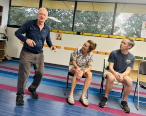An older man stands and gestures while speaking to two seated adults, a woman and a man, in a brightly lit room with colorful floor stripes and large windows. The seated pair are listening attentively to the standing man.