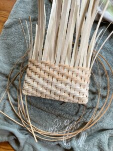 A partially woven basket made of light-colored reeds rests on a gray towel, with loose strips sticking up and several long strands extending outward.