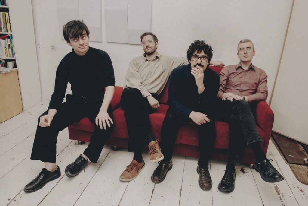 Four men sit casually on a red couch in a white room with wooden floors, looking toward the camera.