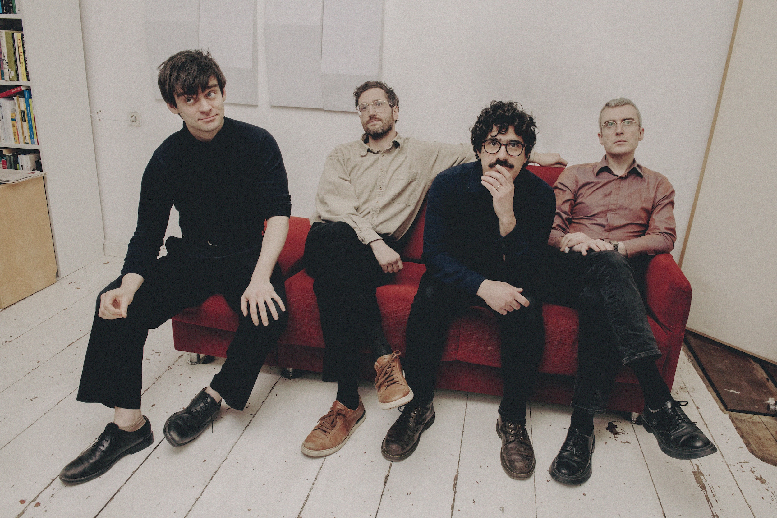 Four men sit casually on a red couch in a white room with wooden floors, looking toward the camera.