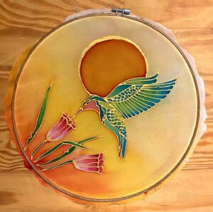 Embroidery art of a colorful hummingbird with blue and green wings drinking from pink flowers, set against a warm yellow and orange background with a large sun. The round hoop frames the artwork on a wooden surface.