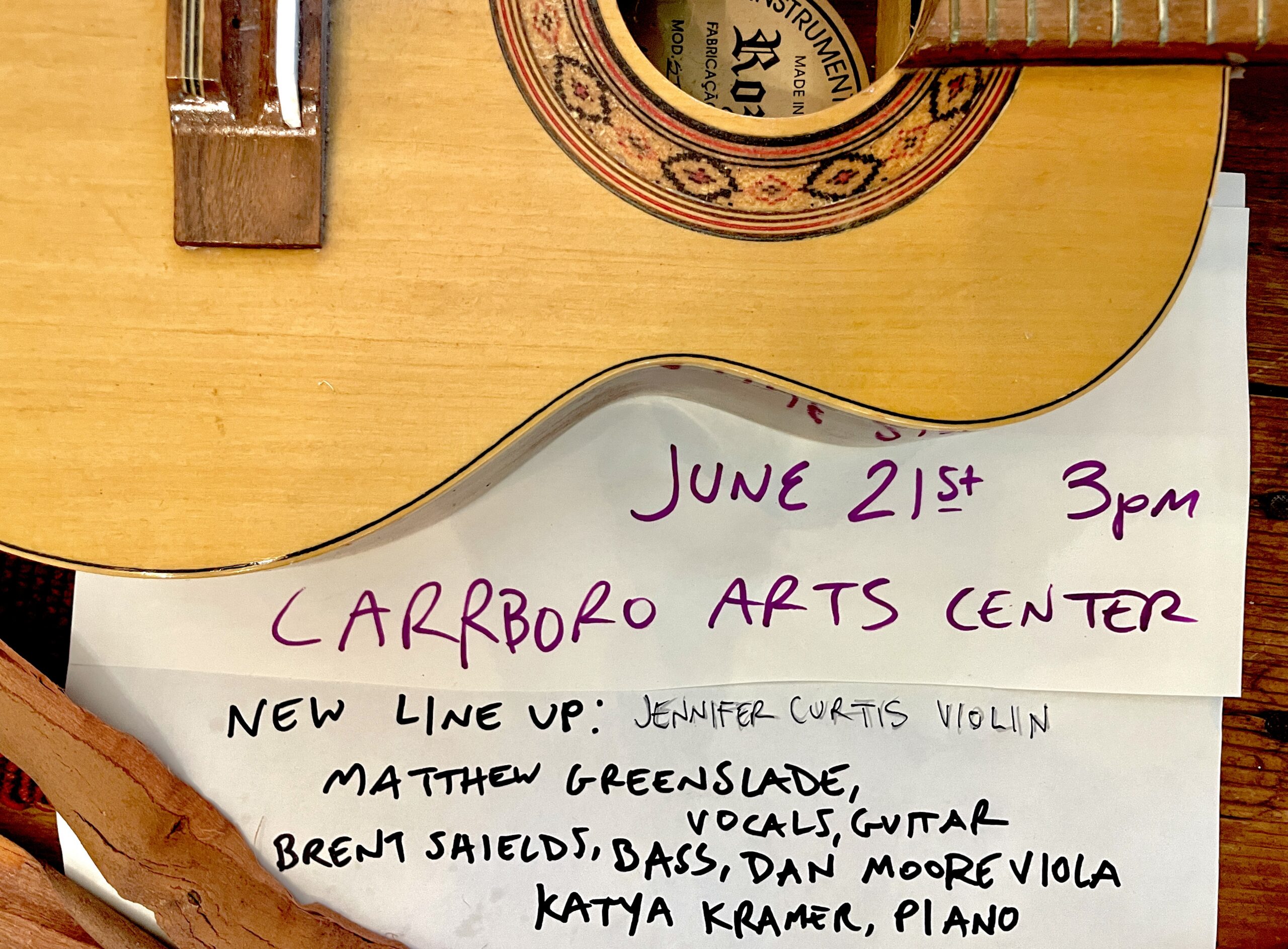 A close-up of a guitar body above a handwritten note announcing a music event: June 21st 3pm, Carrboro Arts Center. Below, a lineup lists names and instruments, including violin, vocals, guitar, bass, viola, and piano.