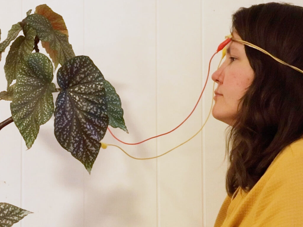 A woman with wires attached to her head faces a plant, with one wire also attached to a leaf.