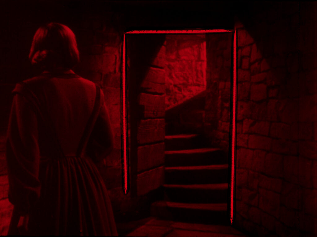 A person stands in a dim, red-lit stone hallway, looking toward stairs leading up through a doorway.