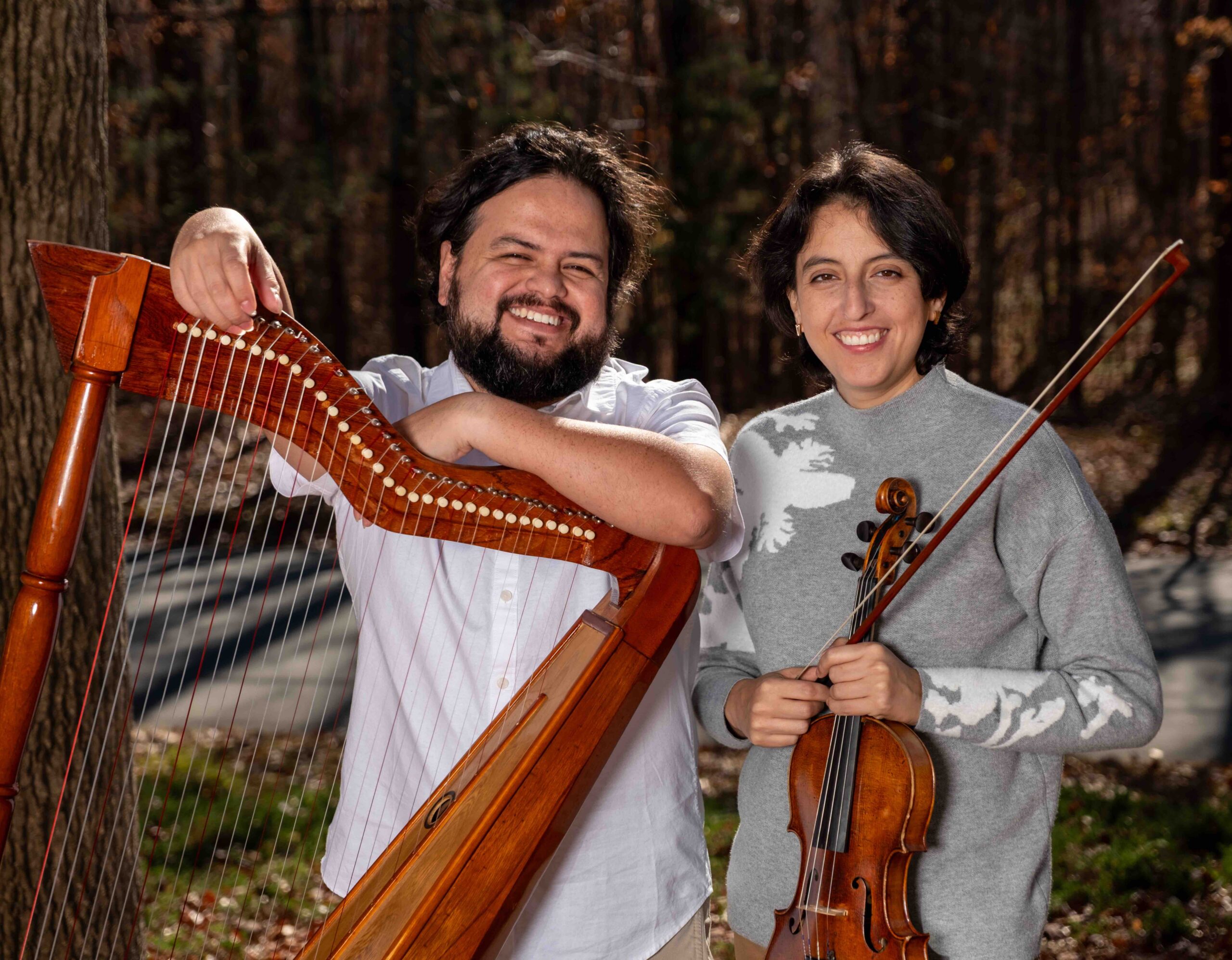 Two people stand outdoors, smiling. One holds a harp, the other holds a violin and bow. Trees and sunlight are in the background.