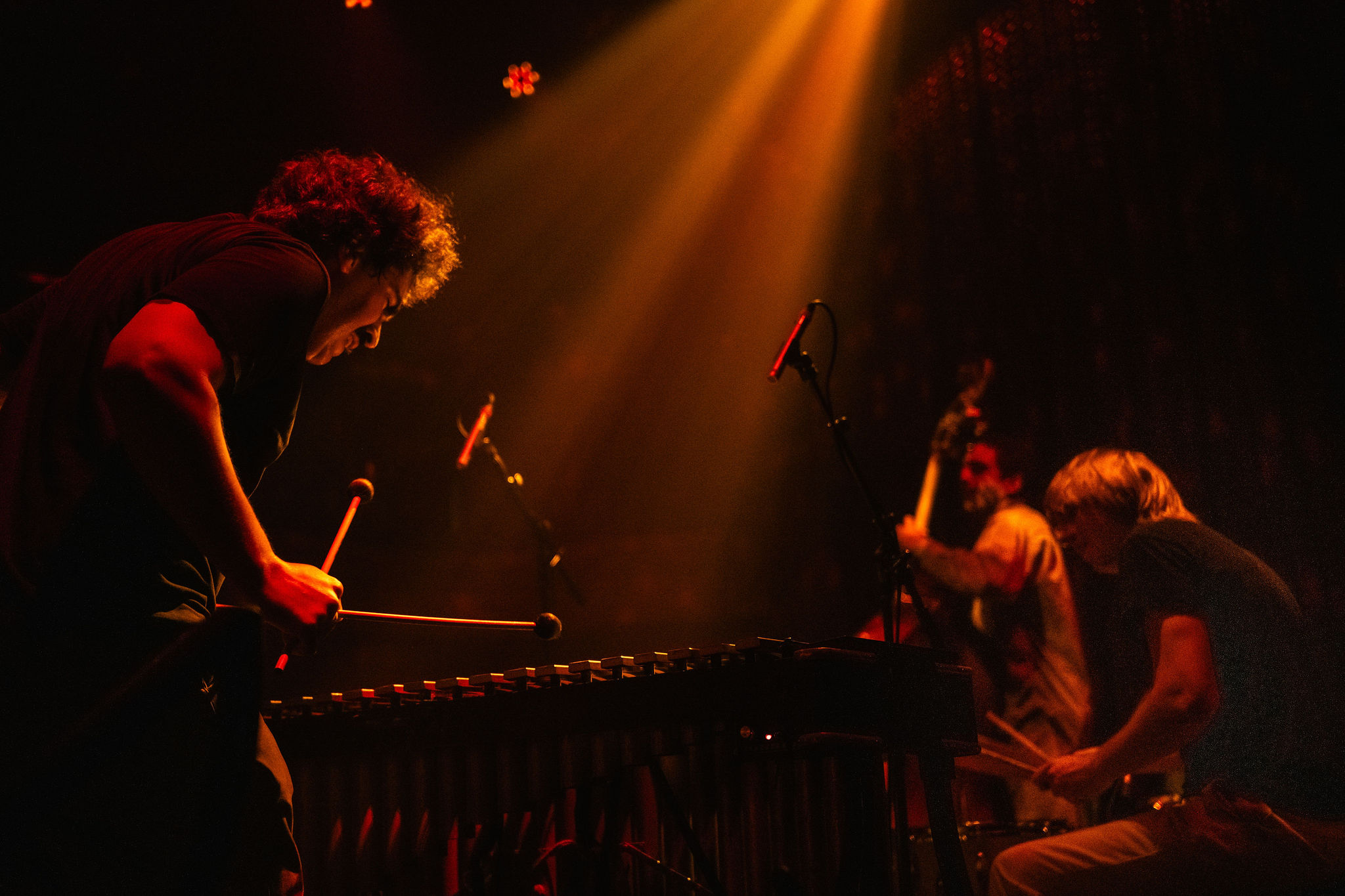 Three musicians play vibraphone, double bass, and drums on a dimly lit stage with orange spotlights.