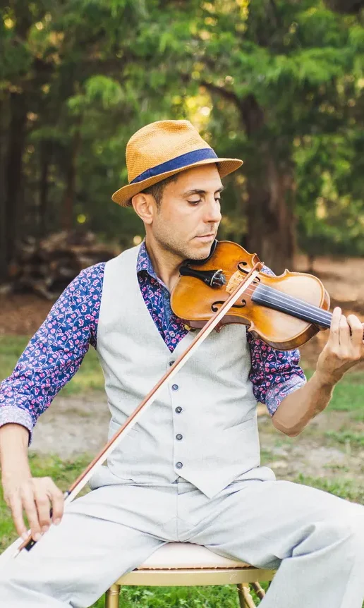 A man in a hat and vest sits outdoors on a chair, playing a violin, with trees in the background.