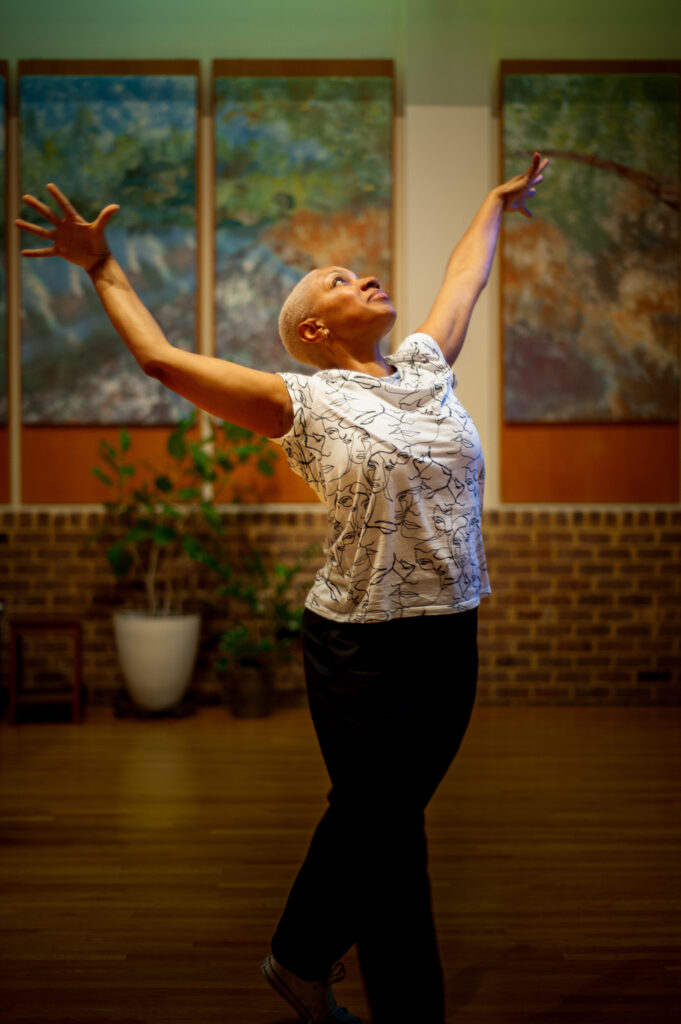 An older woman with short hair dances joyfully indoors, arms raised, with art and plants in the background.