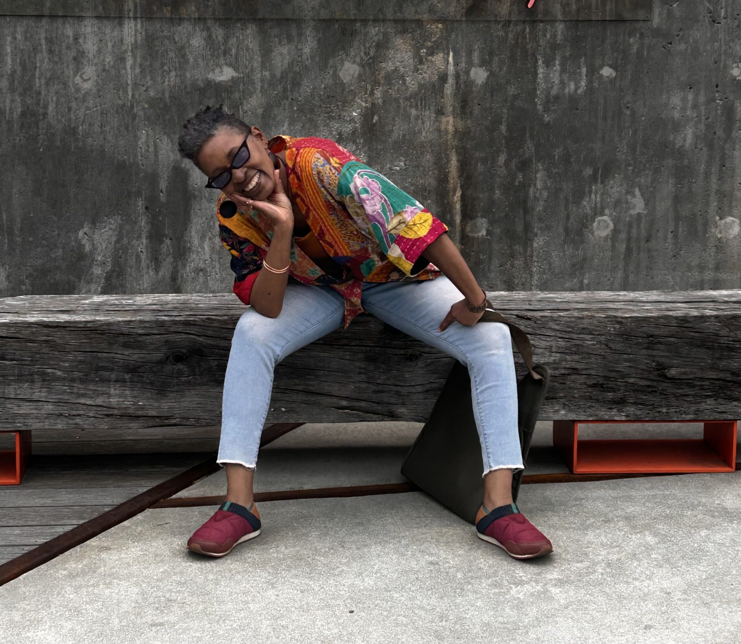 Person in colorful shirt and red shoes sitting on a wooden bench, leaning forward and smiling, with a concrete wall behind.