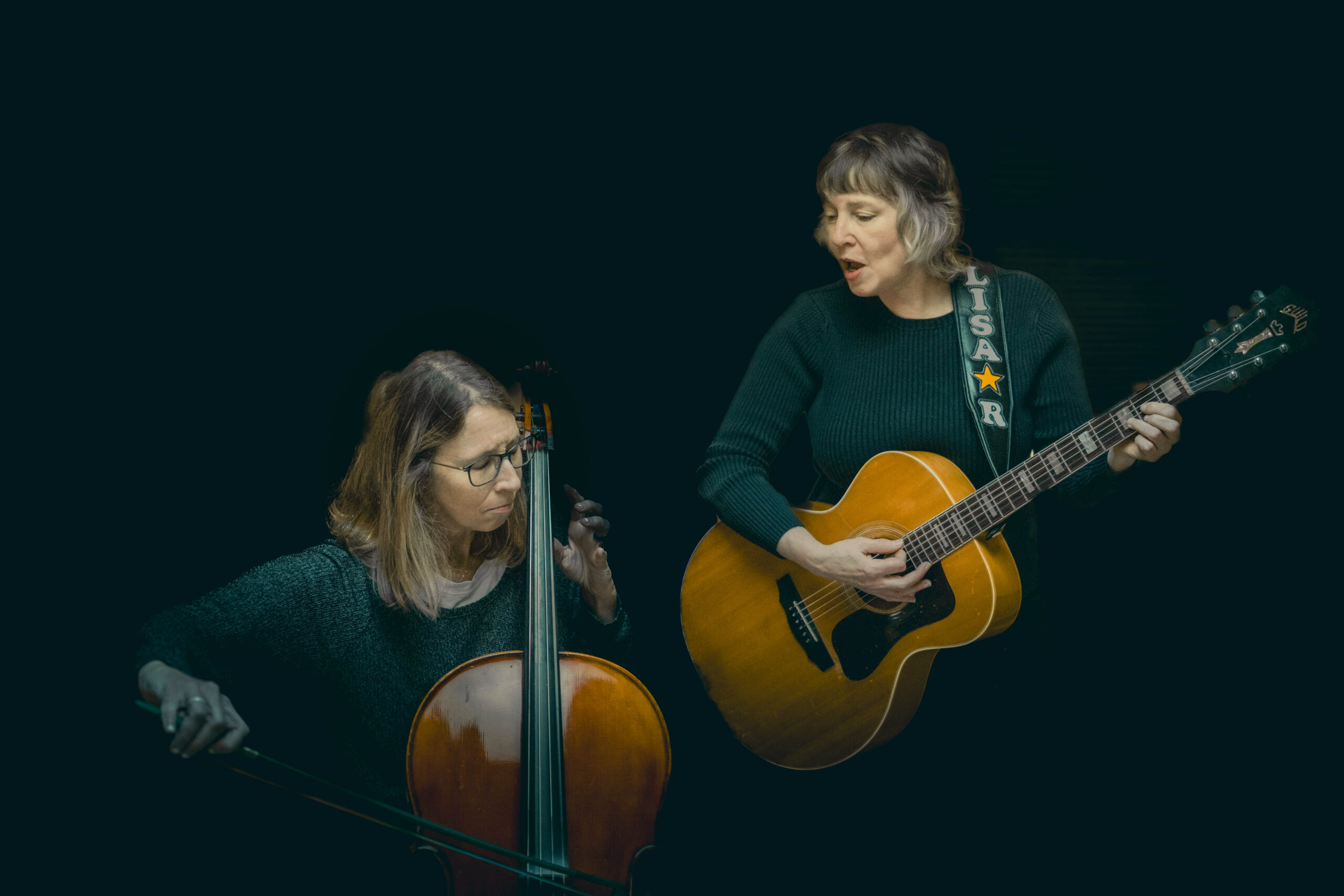 Two women perform: one playing cello, the other singing and playing an acoustic guitar with a name strap that reads LISA R. on it.