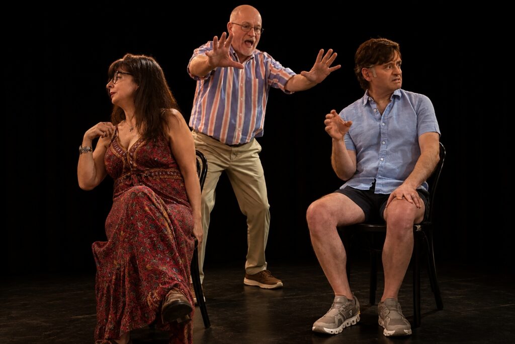 Three adults on stage: a woman and a man sit on chairs, while another man stands behind them gesturing animatedly.