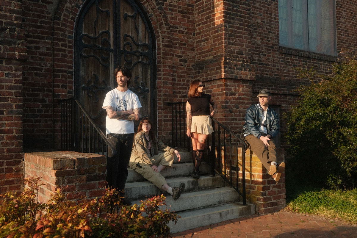 Four people sit and stand on church steps, posing casually in sunlight, with a large wooden door and brick wall behind them.
