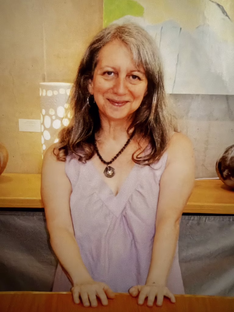 A woman with long gray hair in a purple dress stands smiling in front of a table with pottery and art in the background.