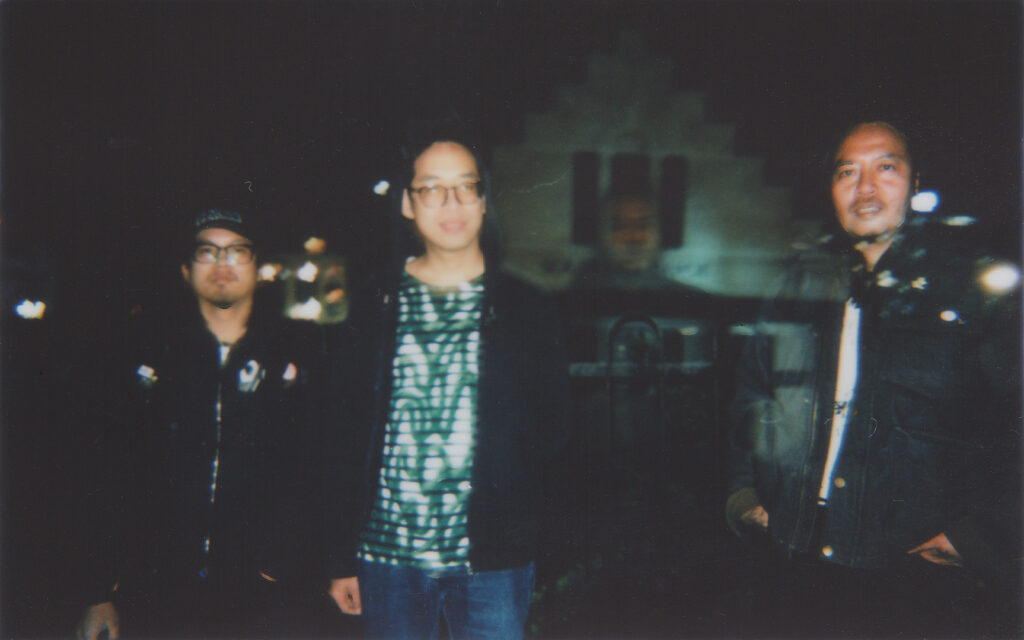 Three men stand outdoors at night, with a blurred, dark background and a faintly visible structure behind them.