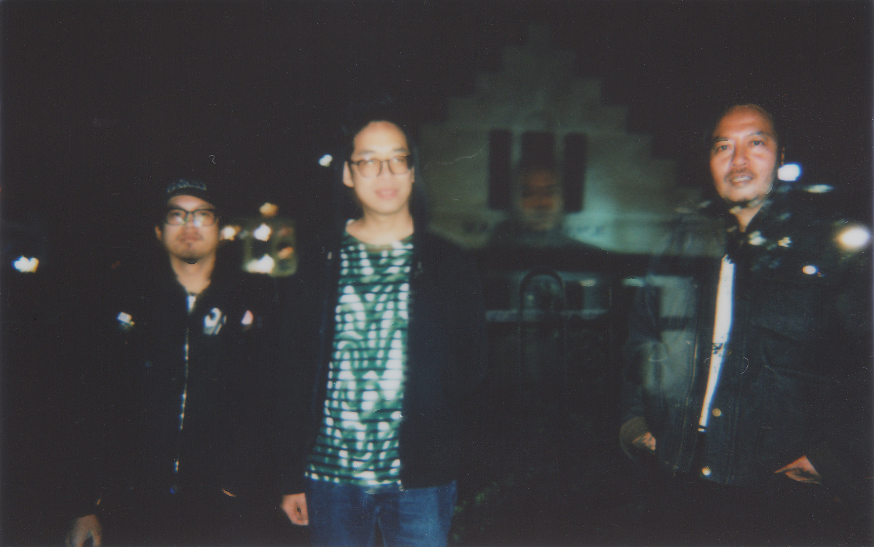 Three men stand outdoors at night, with a blurred, dark background and a faintly visible structure behind them.