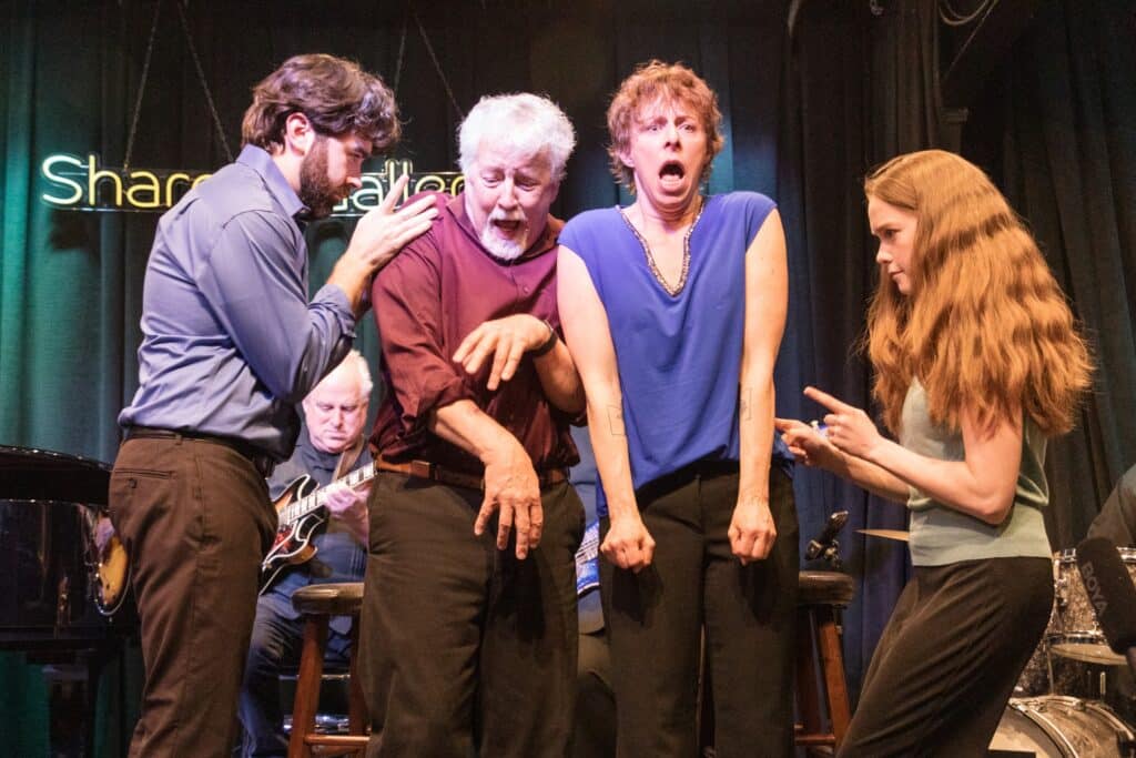 Four people on stage express intense emotions; musicians play in the background under a neon sign and stage lights.