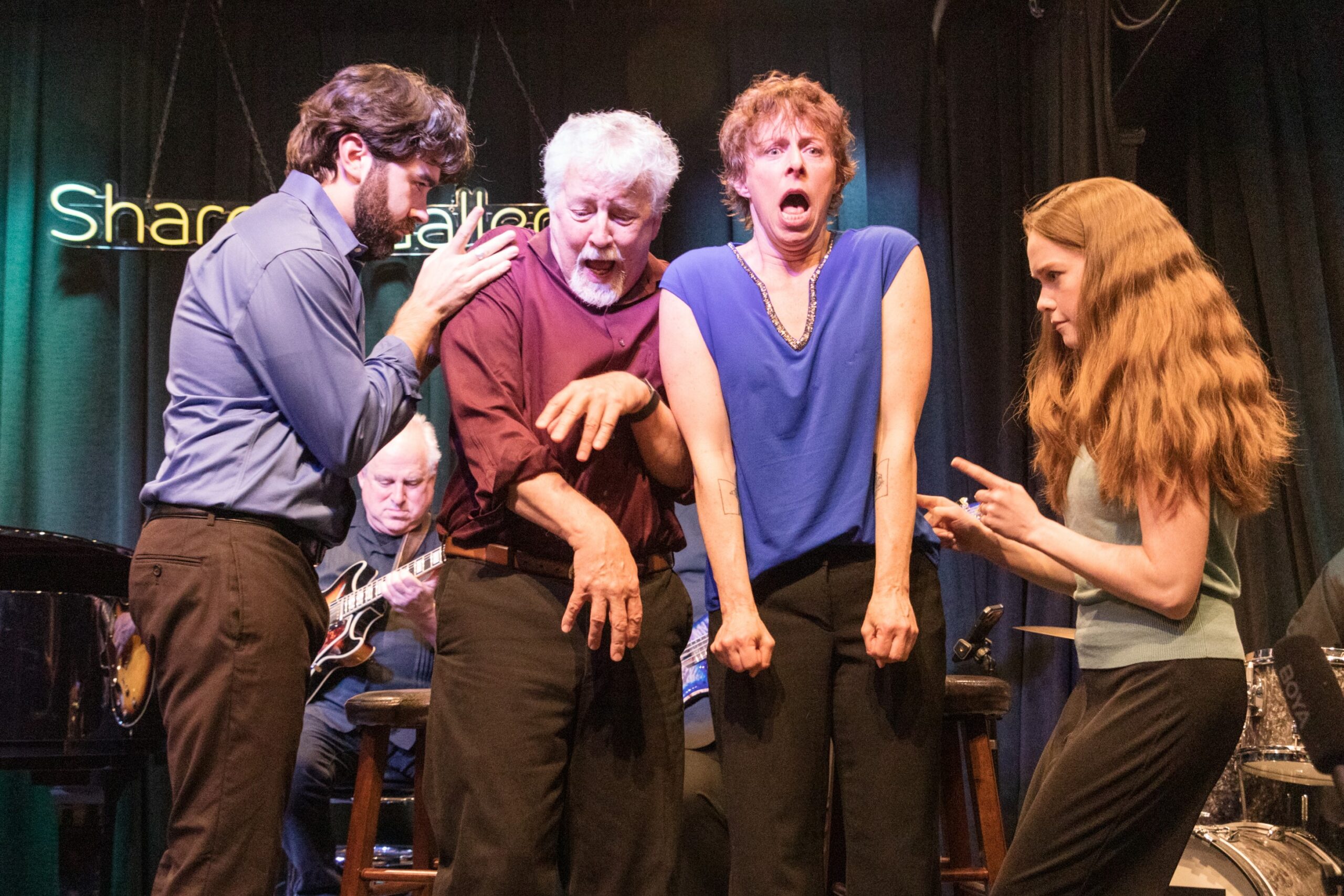 Four people on stage express intense emotions; musicians play in the background under a neon sign and stage lights.