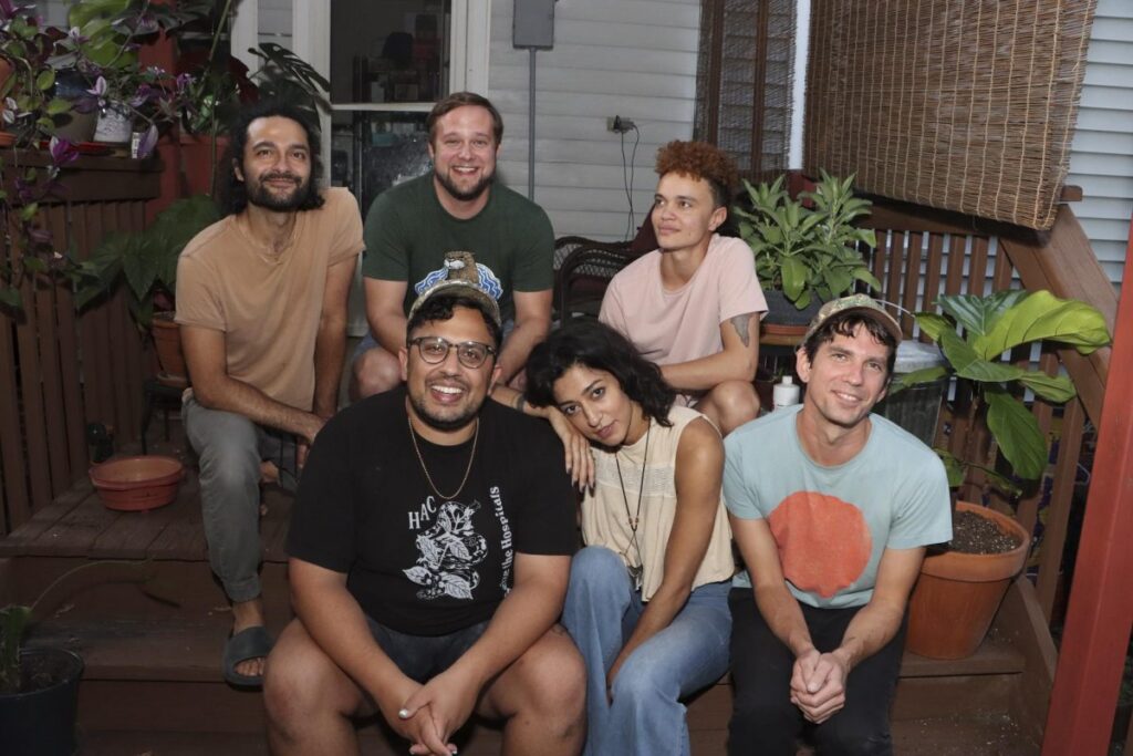 Six people sit and smile on a porch surrounded by potted plants, with a house and window in the background.