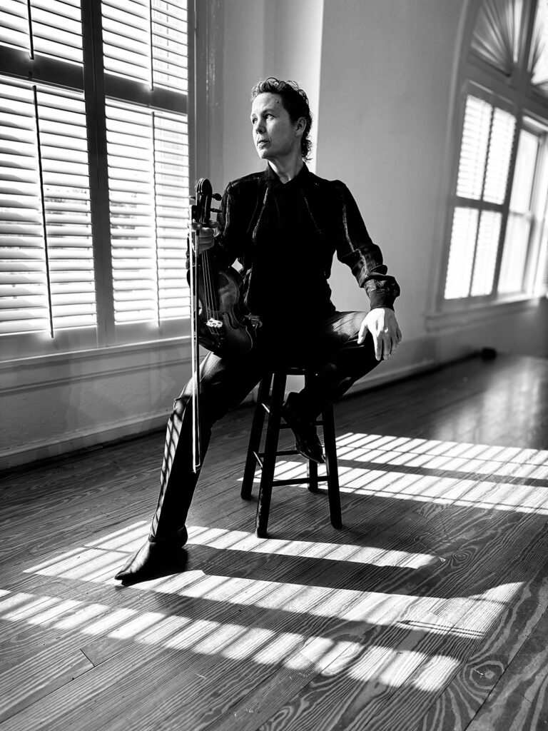 A person sits on a stool holding a violin in a sunlit room with large windows and wooden floors.