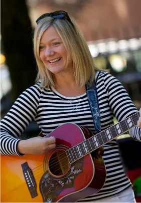 Smiling woman in a black-and-white striped shirt playing an acoustic guitar outdoors.