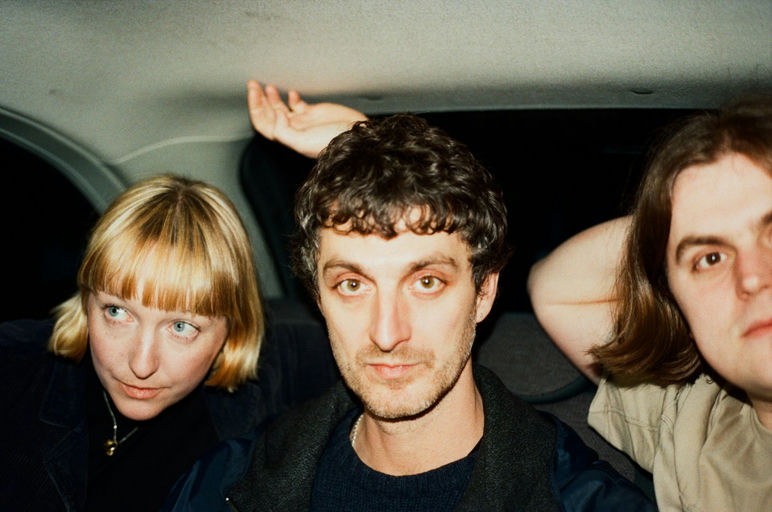 Three people sit closely together in the backseat of a car, looking toward the camera with neutral expressions.