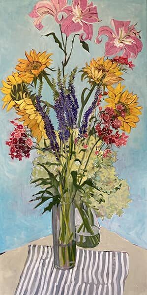 A glass vase with sunflowers, pink lilies, red, purple, and white flowers on a striped cloth against a blue background.