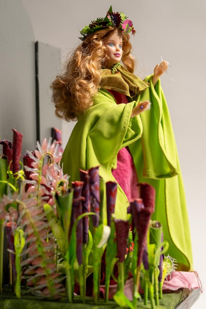 A doll with long red hair wearing a green cloak and floral crown stands behind a collection of small, colorful model pitcher plants.