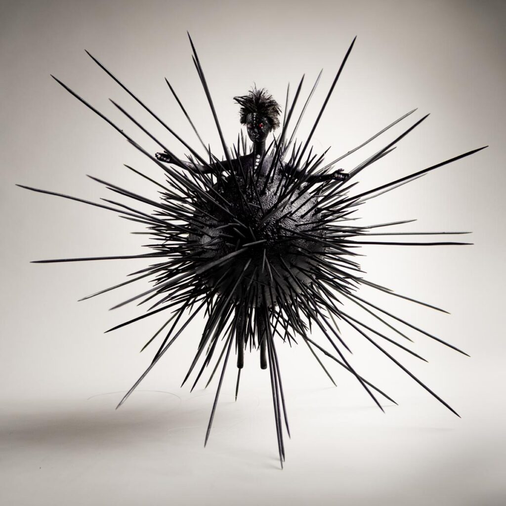 A figure with a spiky head emerging from a large, black, sphere-shaped sculpture covered in long, sharp needles.