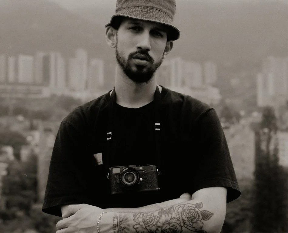 A bearded man wearing a bucket hat and a black t-shirt, with a camera hanging around his neck and rose tattoos on his arm.