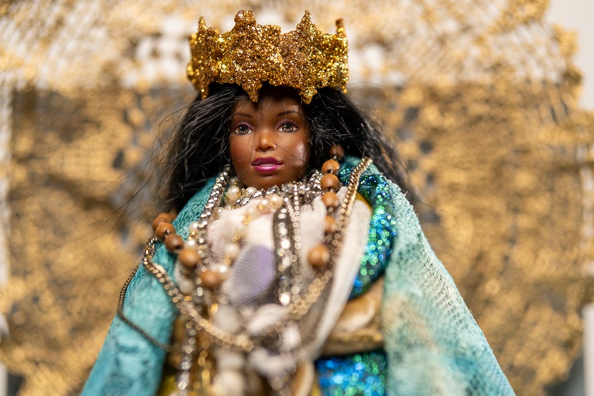A doll dressed in a teal cloak and a gold crown, adorned with multiple necklaces.