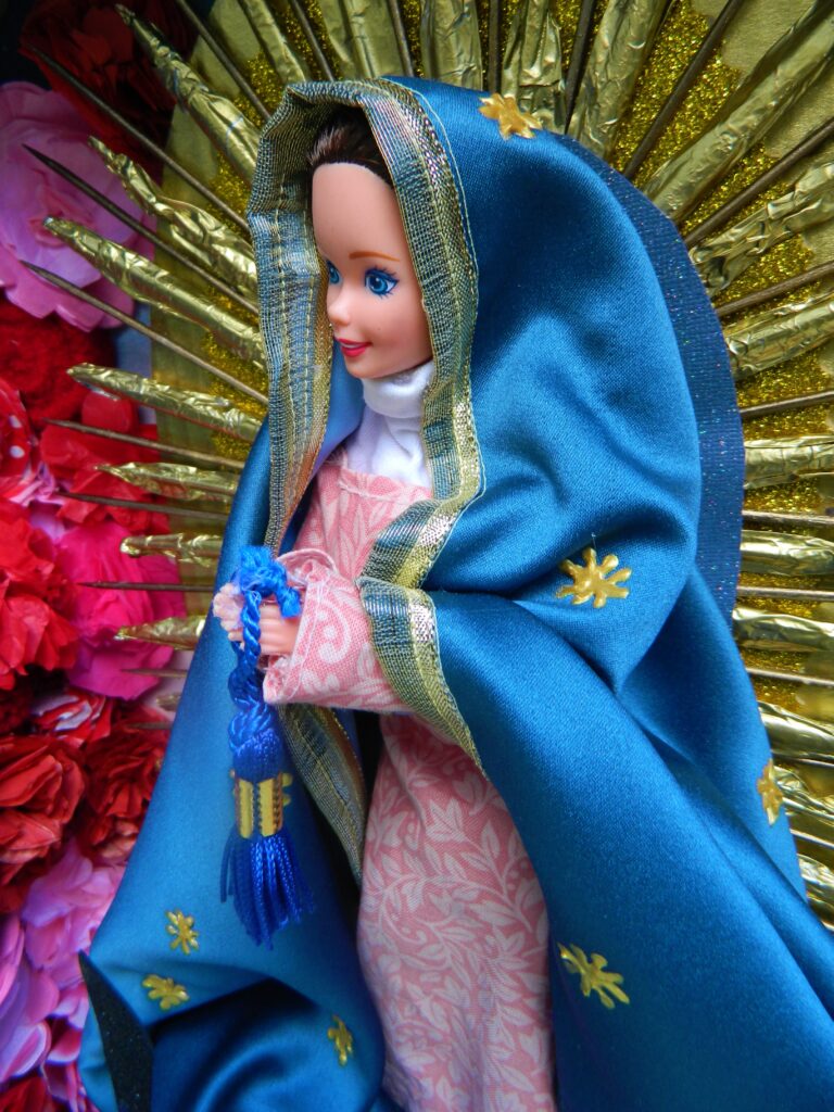 A doll dressed as the Virgin of Guadalupe, wearing a blue mantle and pink gown in front of a golden sunburst.