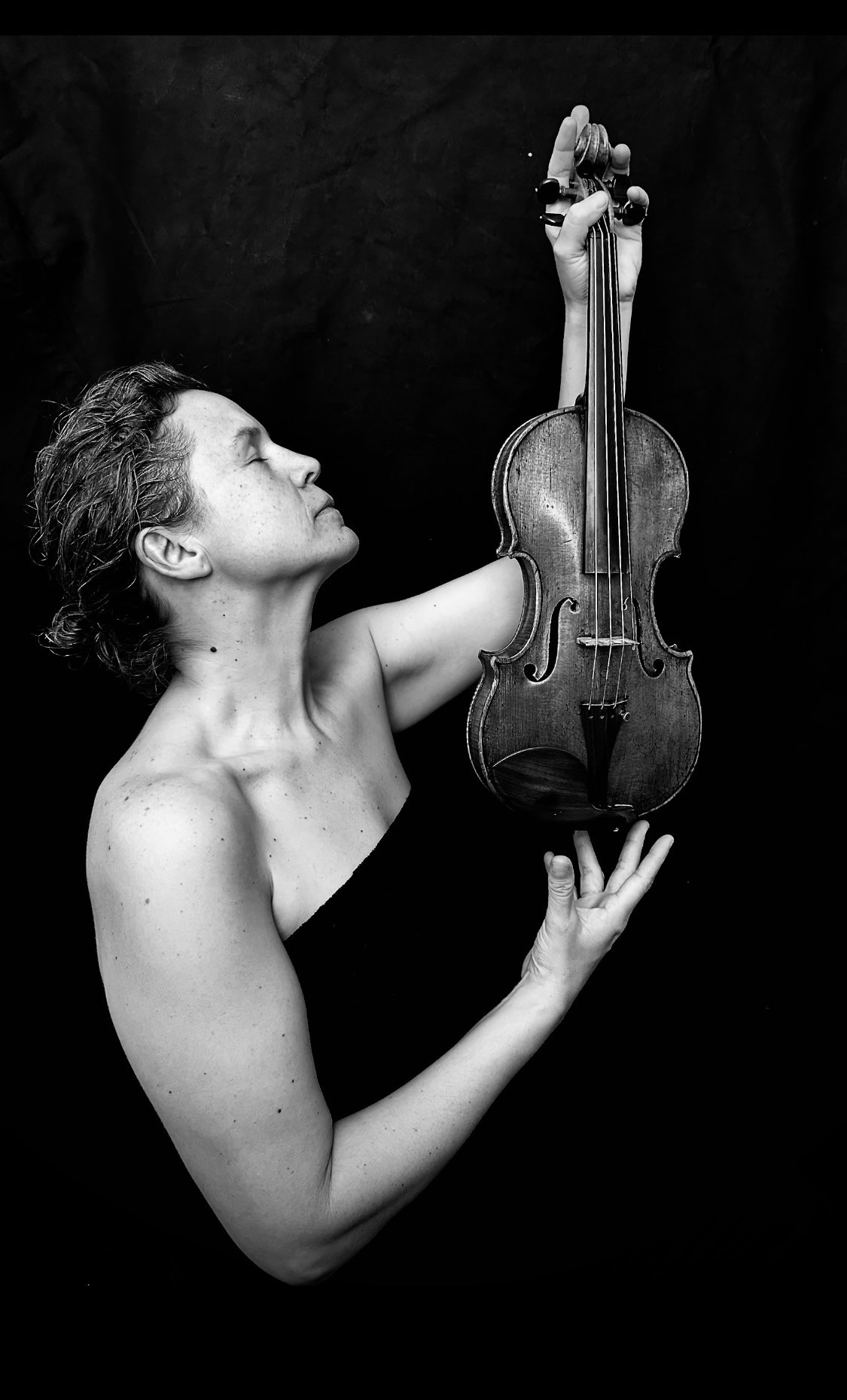 A woman with eyes closed holds a violin vertically in front of her.